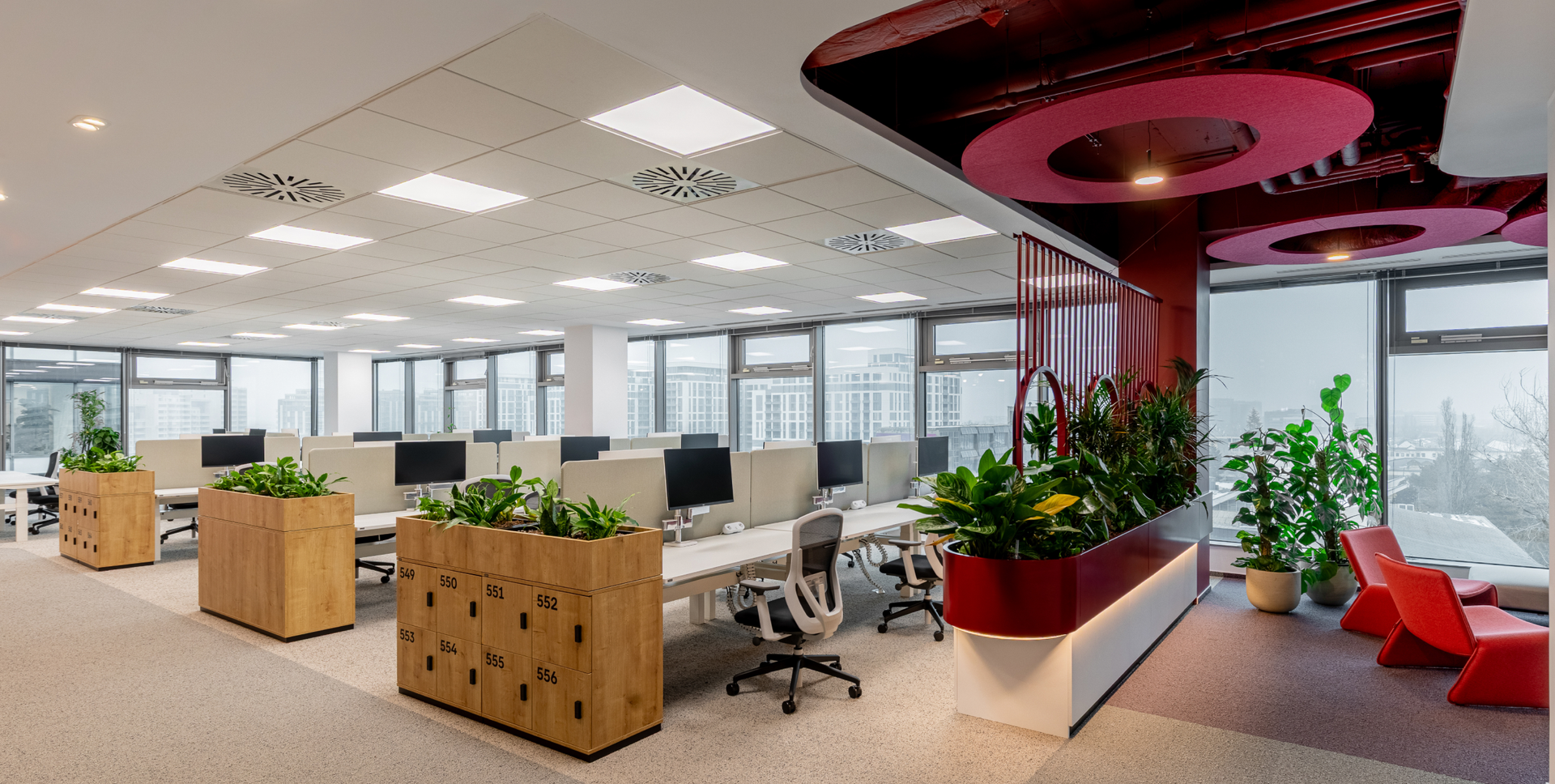 Modern open-plan office with desks, plants, large windows, and a lounge area with red accents.