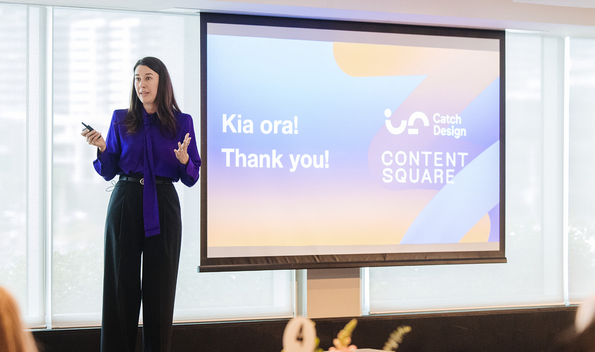 A woman in a purple blouse presents, holding a remote, next to a screen displaying "Kia ora! Thank you!" and company logos.