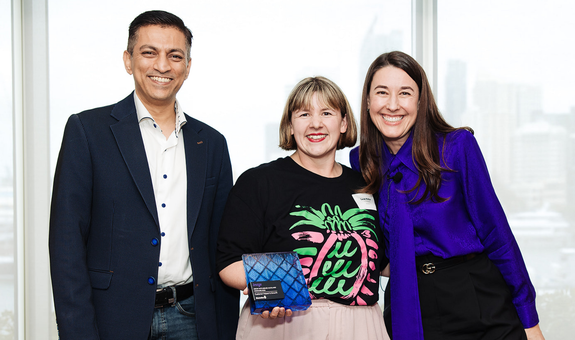 A man and two women smiling, with one woman holding a blue award, in front of a city view.