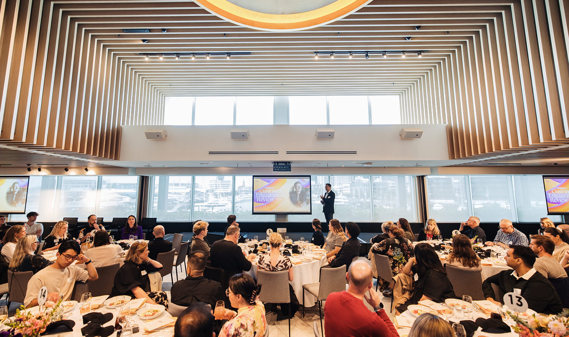 A speaker presents to an audience seated at round tables in a modern event hall with large screens.