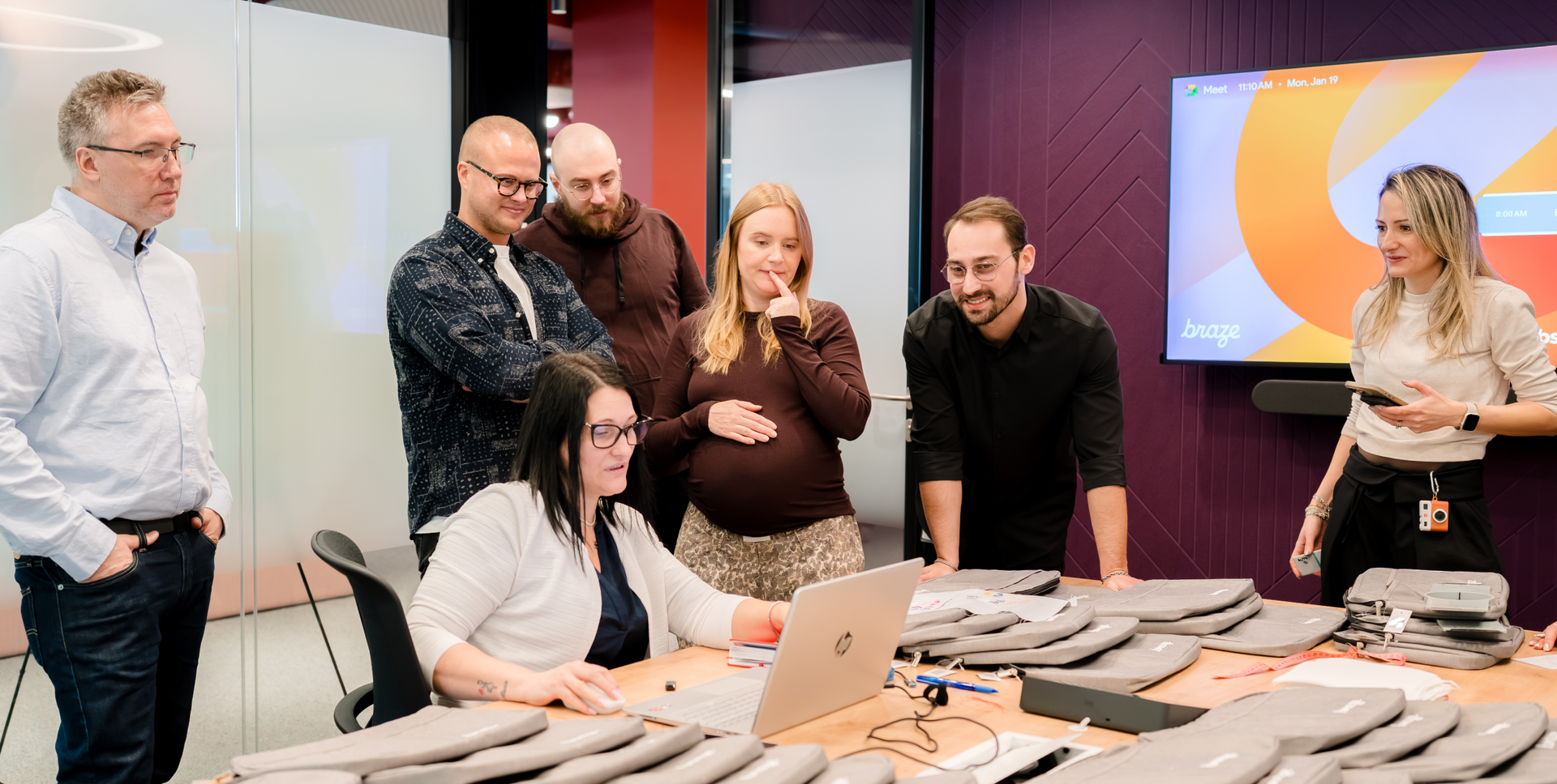 A group of seven people collaborating around a laptop in a modern office, with tech accessories on the table.