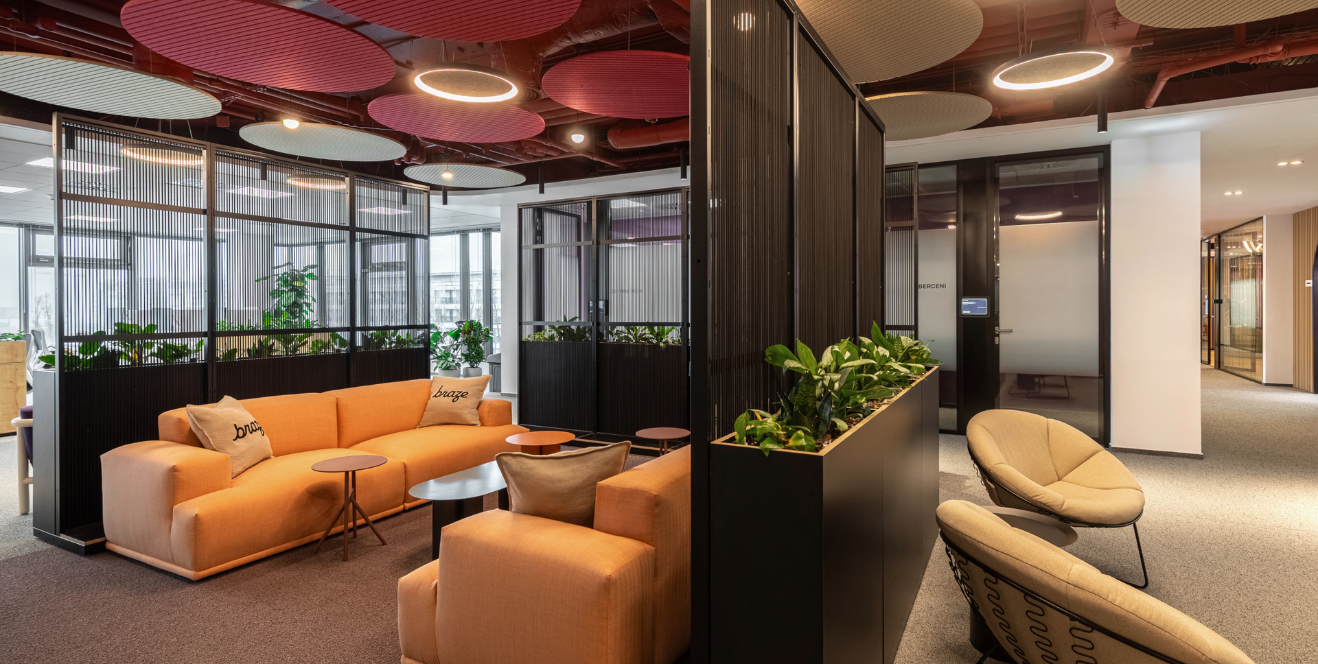 A modern office lounge with orange sofas, green plants in dividers, and circular acoustic ceiling panels.