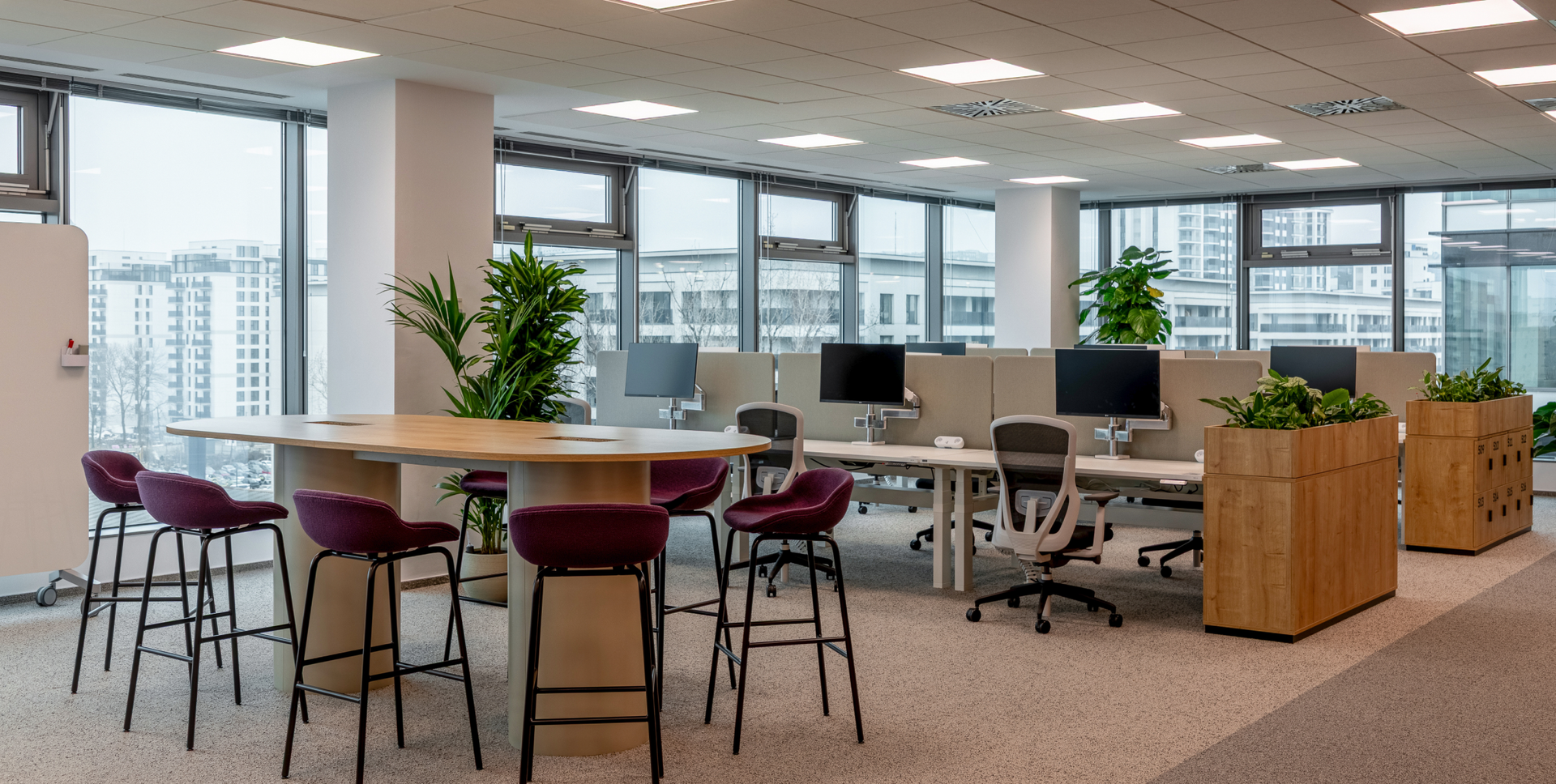 A modern open-plan office featuring a collaborative high-top table with stools, individual workstations, and large windows with a city view.