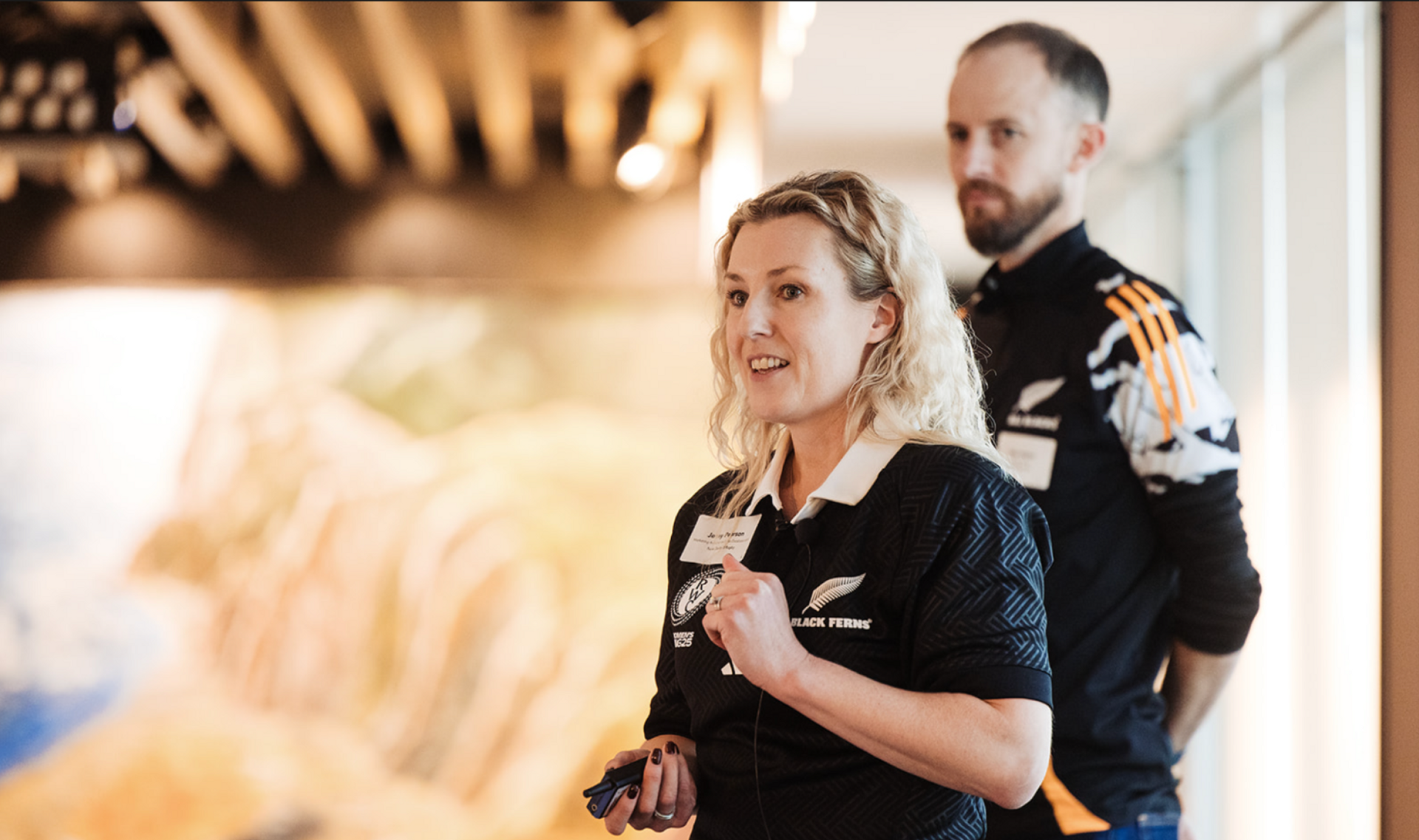 A blonde woman in a black Black Ferns polo shirt speaks, with a bearded man in an Adidas shirt standing behind her.