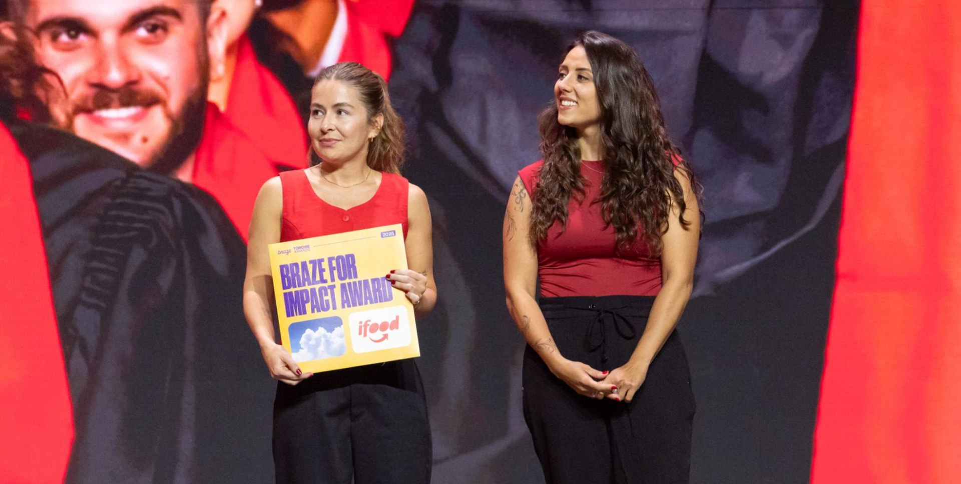 Two women on a stage, one holding a yellow "BRAZE FOR IMPACT AWARD" plaque with the iFood logo.
