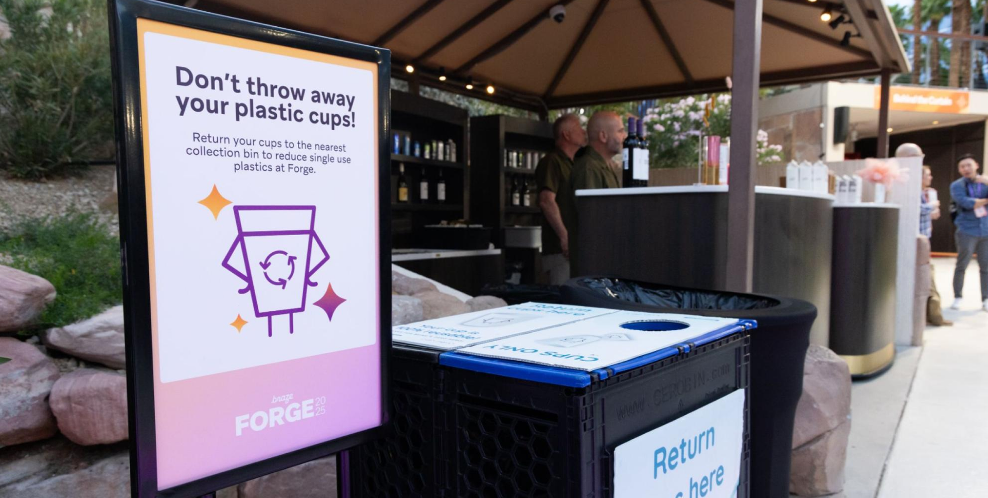 A sign at a Forge event promoting plastic cup recycling, with a collection bin and beverage counter in the background.