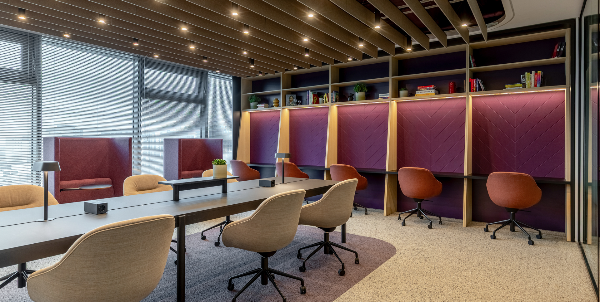 A vibrant, modern co-working space with a long central table, window-side booth seating, and a wall of individual purple-paneled desks with bookshelves.