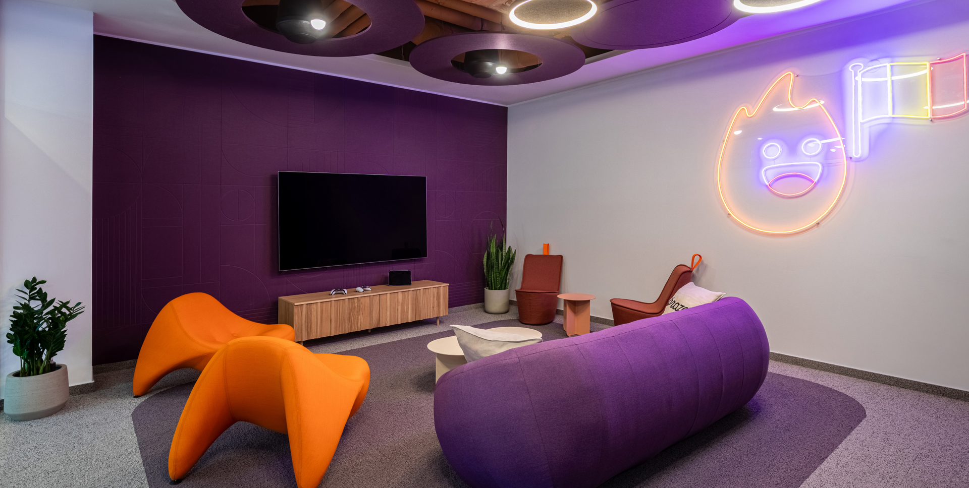 A vibrant lounge area with a purple wall, orange chairs, a purple sofa, a large TV, and a neon flame character sign.