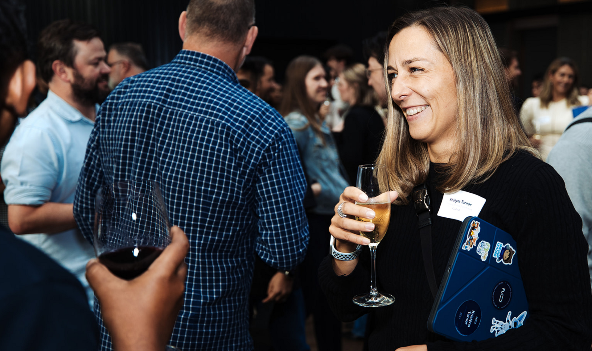 A woman smiles, holding a glass of champagne and a tablet, at a crowded event.