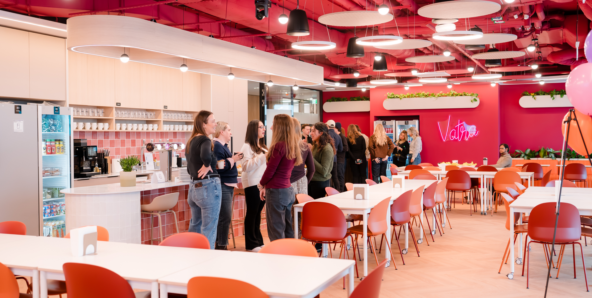 A modern, vibrant office cafeteria with pink walls, orange chairs, a kitchenette, and people socializing.