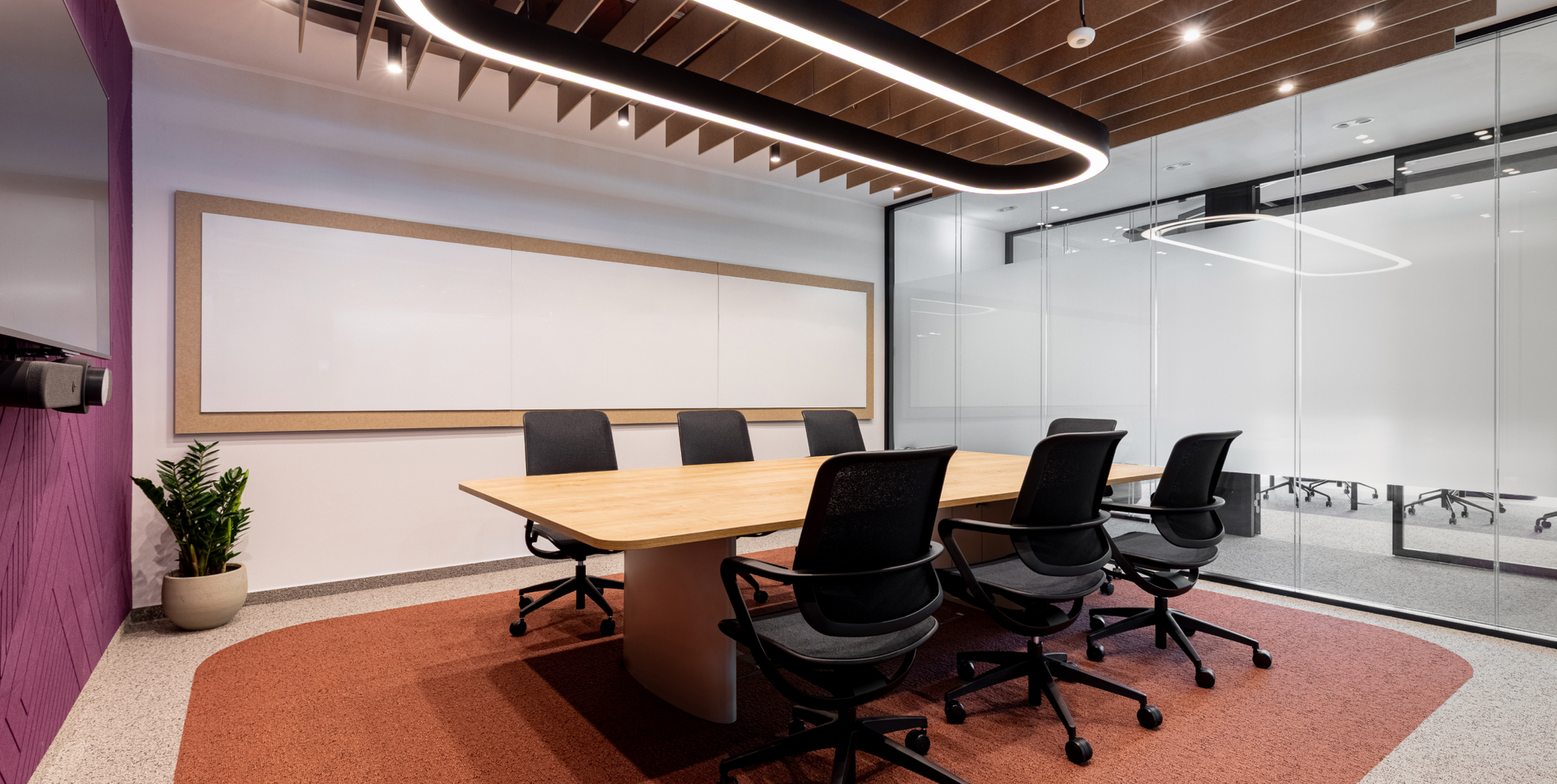A modern meeting room with a light wood conference table, black chairs, large whiteboard, and purple and terracotta accents.